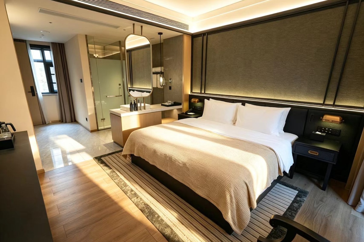 A modern hotel room with a large bed, a bedside table, a nightstand, a mirror, and a bathroom with a shower and a toilet.