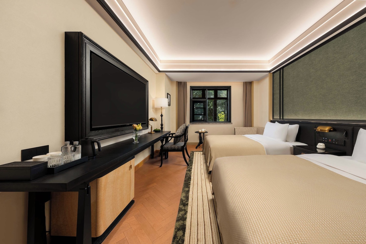 A hotel room with a large flat-screen TV, a desk with a lamp, two beds, and a window with curtains.