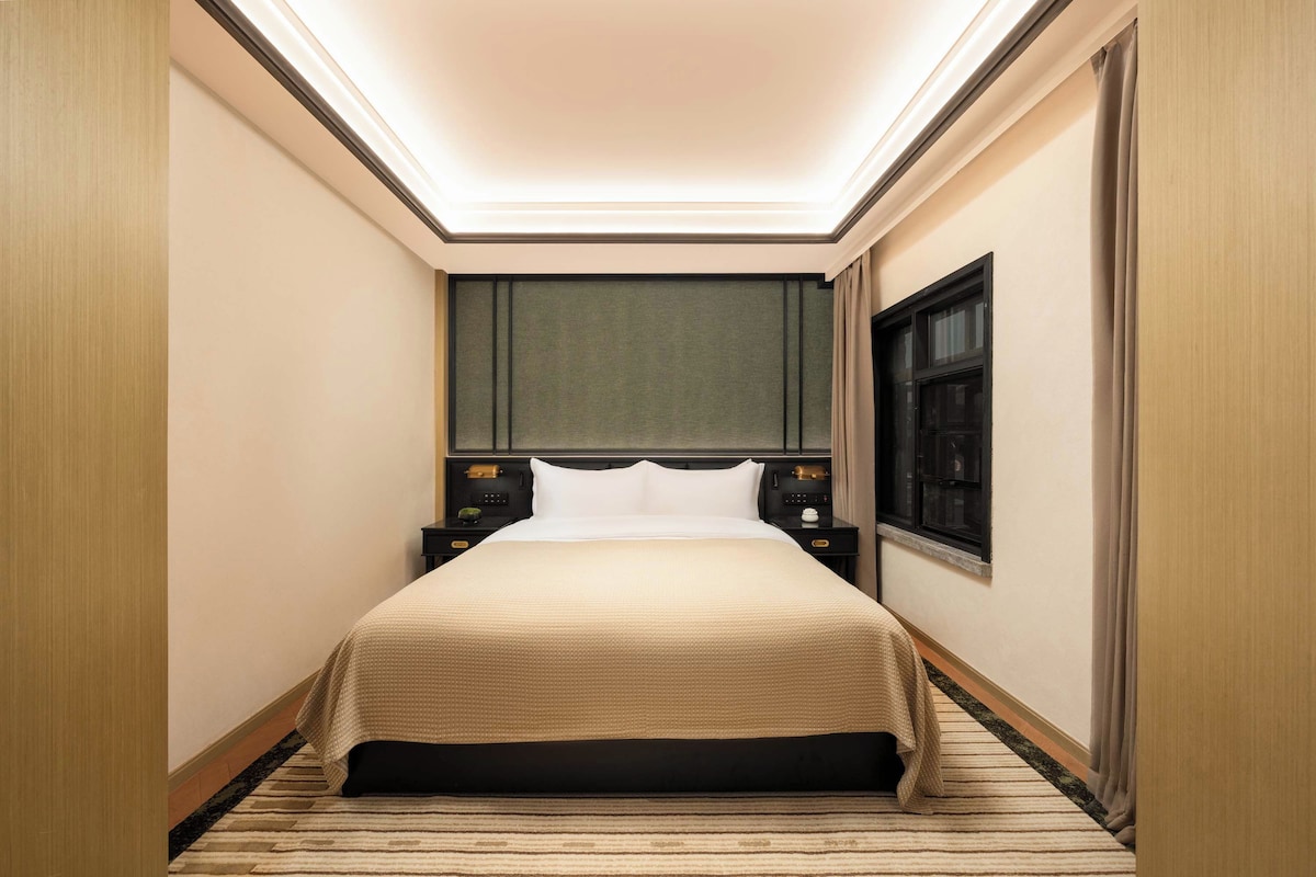 A hotel room with a bed, bedside tables, a window, and a ceiling light.
