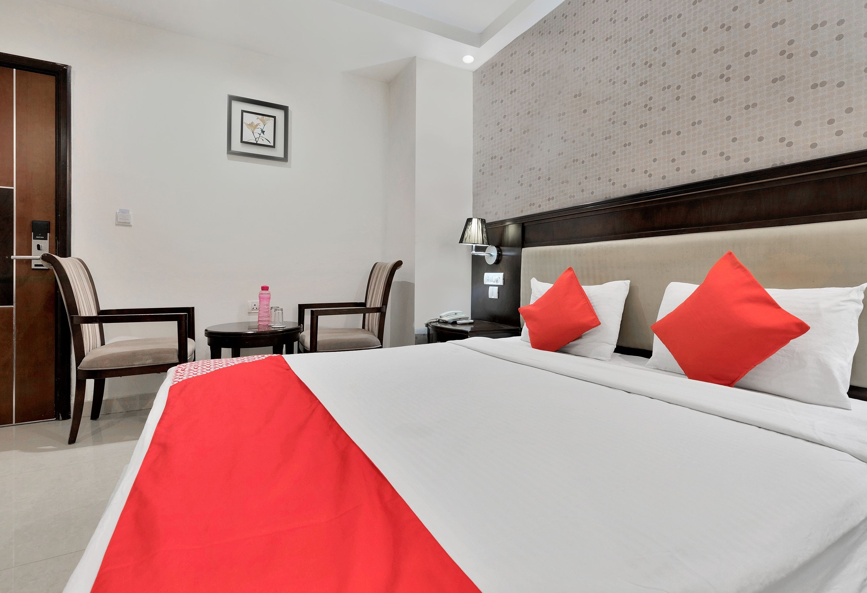 Quarto de hotel com cama, duas cadeiras, mesinha, criado-mudo e luminária na parede.