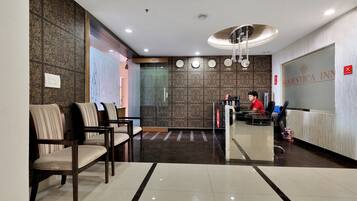 Área de recepção de hotel com recepcionista, relógio e placa de 'Não Fumar'.