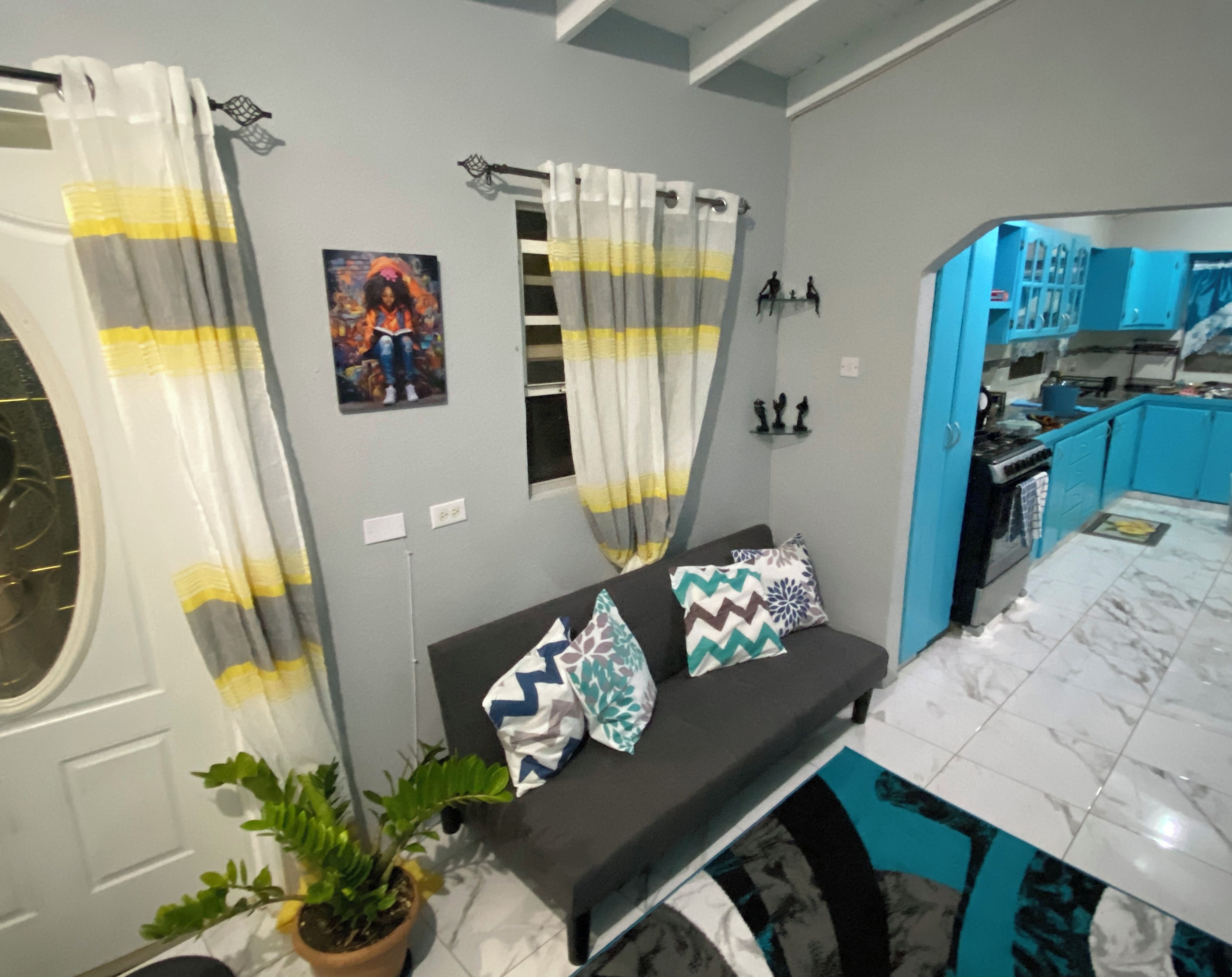 A modern kitchen with blue cabinets, a dining area with a black sofa and patterned cushions, and a grey wall with a framed picture.