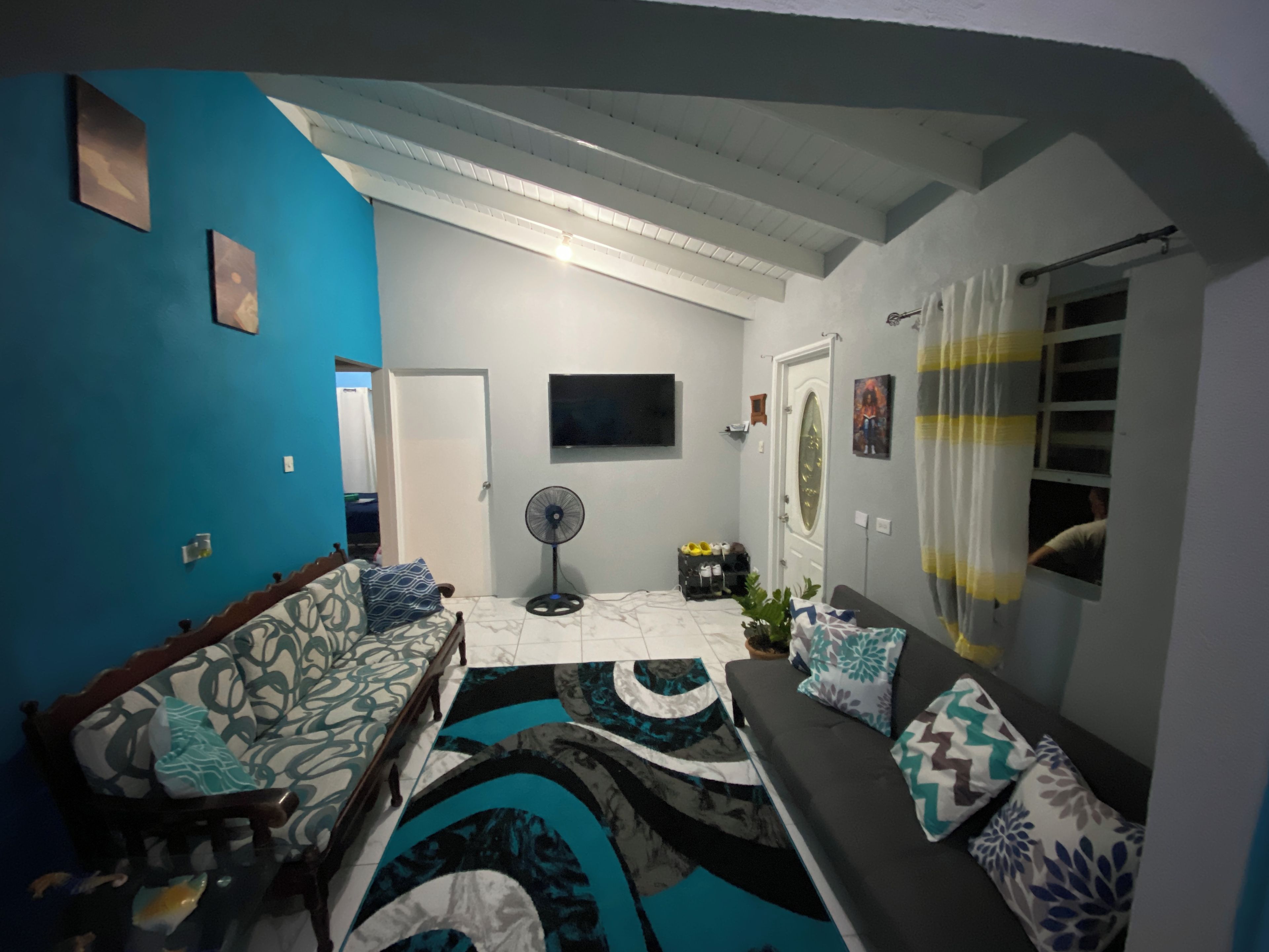 A living room with a sofa, a chair, a TV, a fan, and a door leading to another room.