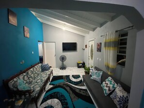 Uma sala de estar com sofá, poltrona, televisão, ventilador e porta que dá para outro cômodo.