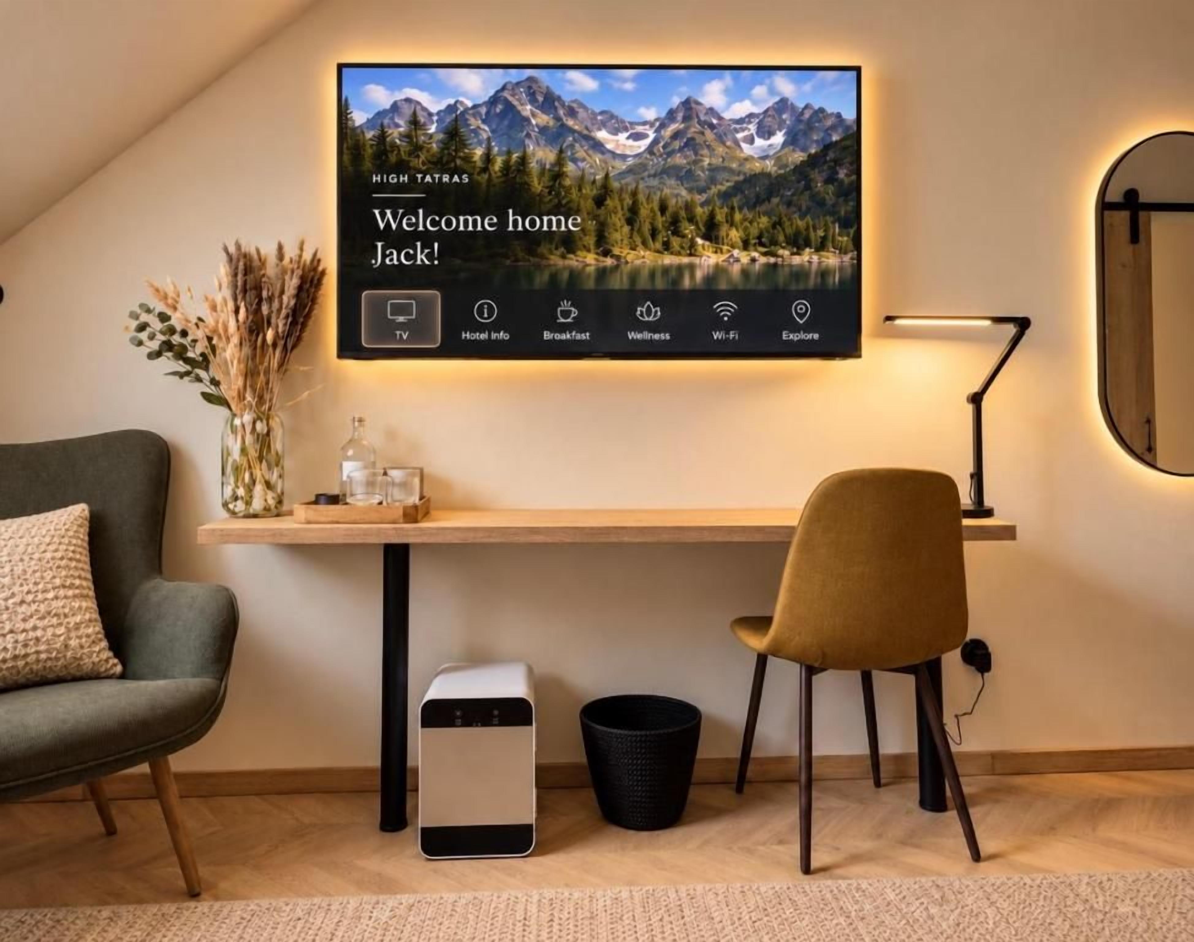 A modern hotel room with a large TV displaying a mountain landscape, a desk with a chair, a small trash bin, and a vase with dried flowers.