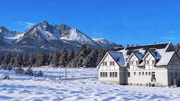 A snow-covered landscape with a traditional chalet-style building in the foreground and a mountain range in the background.