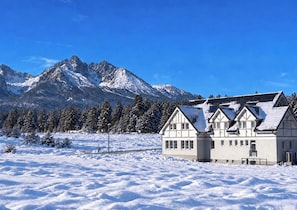 Paisagem coberta de neve com um edifício tradicional de chalé em primeiro plano e uma cordilheira ao fundo.