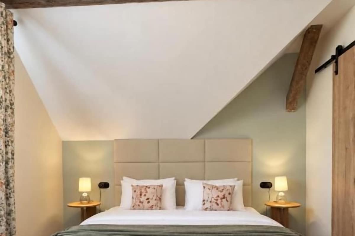 Een slaapkamer met een groot bed, houten balken, twee bedlampen en een visgraat vloerpatroon.