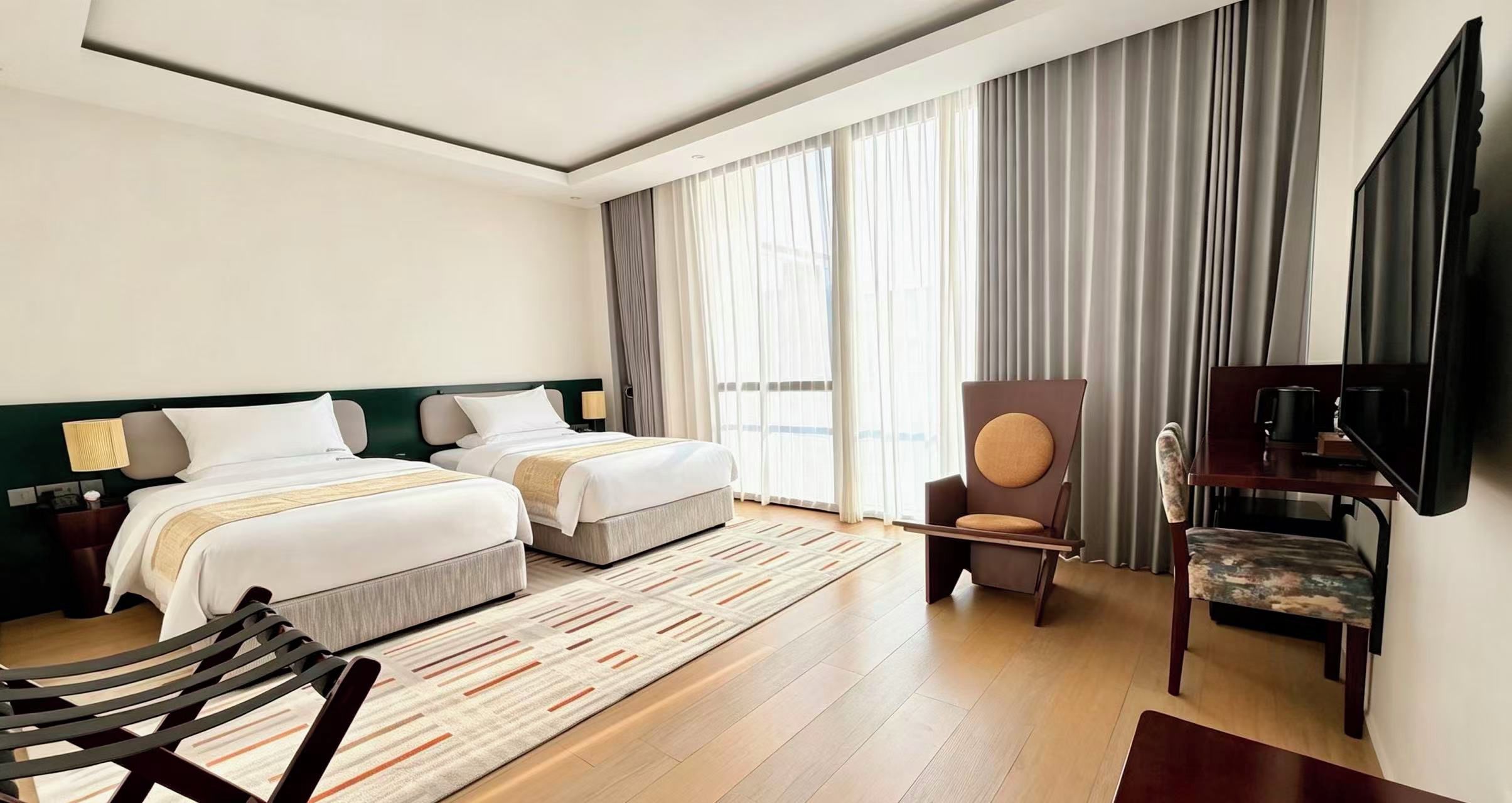Habitación de hotel con dos camas, televisión, una silla y un ventanal con cortinas.