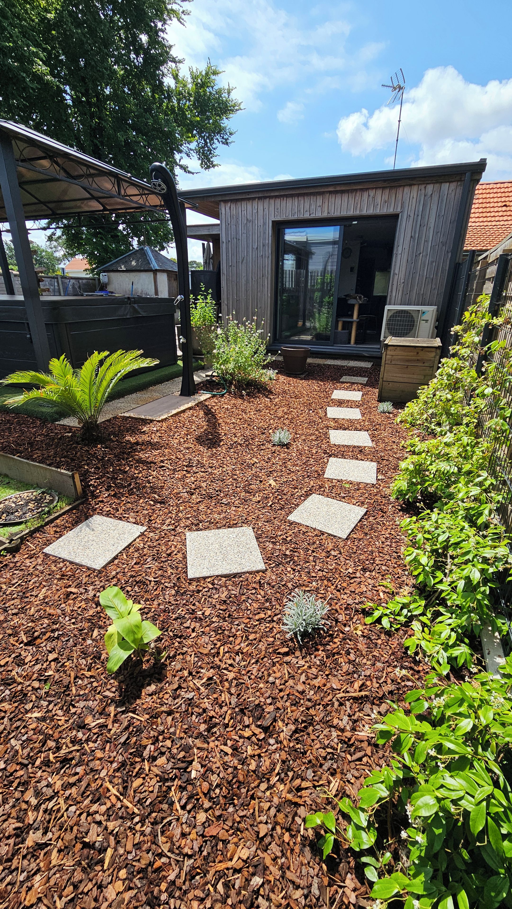 Un sendero en un jardín conduce a un edificio moderno con puerta de vidrio, rodeado de plantas variadas y una cerca de metal.