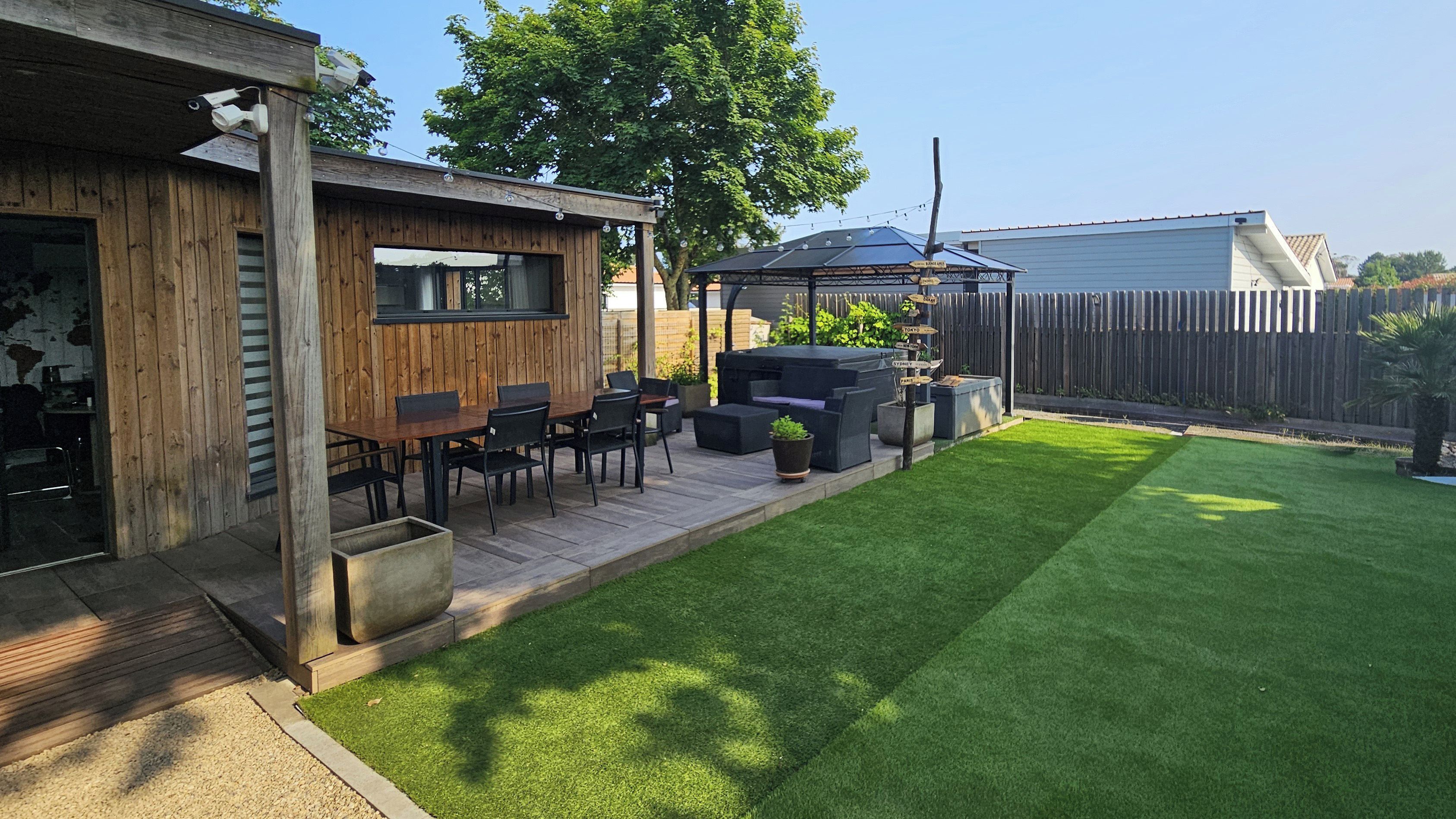 Un patio trasero con terraza de madera, mobiliario exterior, zona de barbacoa y césped cuidado.