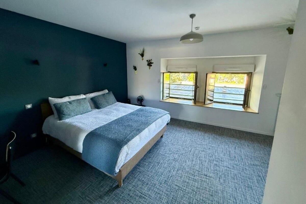 A bedroom with a bed, a chair, a desk, and two windows with a view.