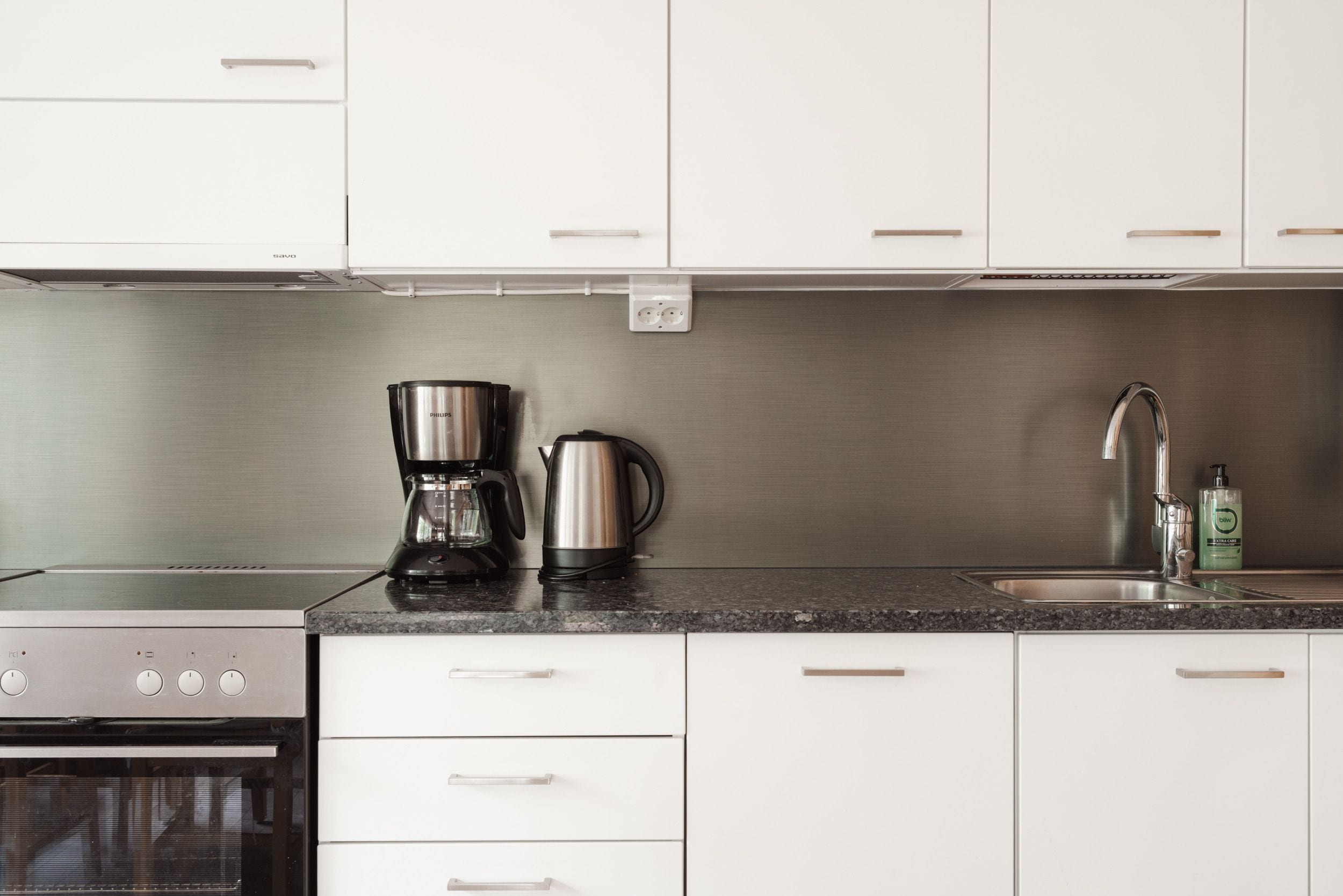 Une cuisine moderne avec des meubles blancs, une cuisinière en acier inoxydable, une machine à café, une bouilloire et un évier avec un robinet.