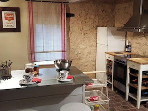 A kitchen with a stone wall, a refrigerator, an oven, and a dining table with a bowl and cups. - Vrbo Property (Onlay)