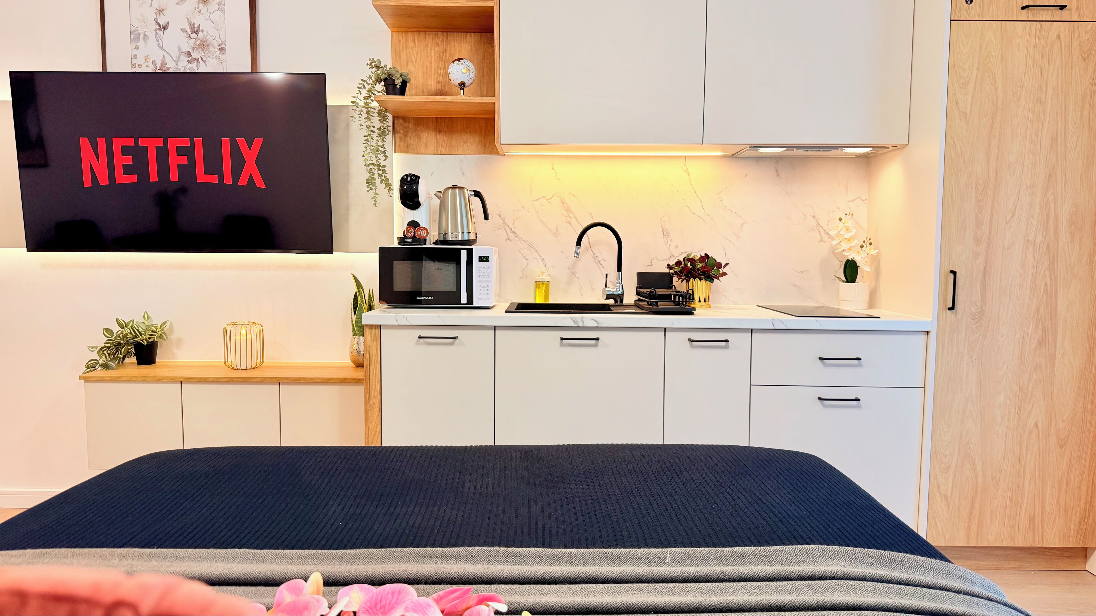 A modern bedroom with a bed, a TV displaying 'NETFLIX,' a kitchen area with a microwave, and a sink.