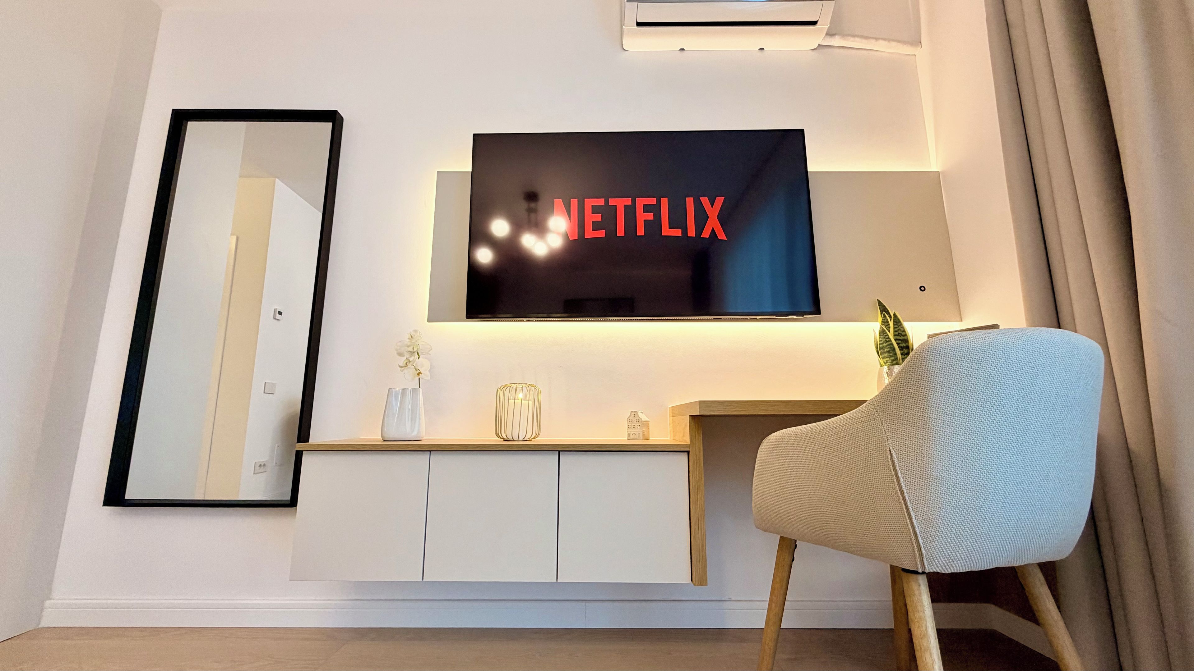 Una sala moderna con un televisor de pantalla plana montado en un mueble blanco, un espejo grande y un sillón cómodo.