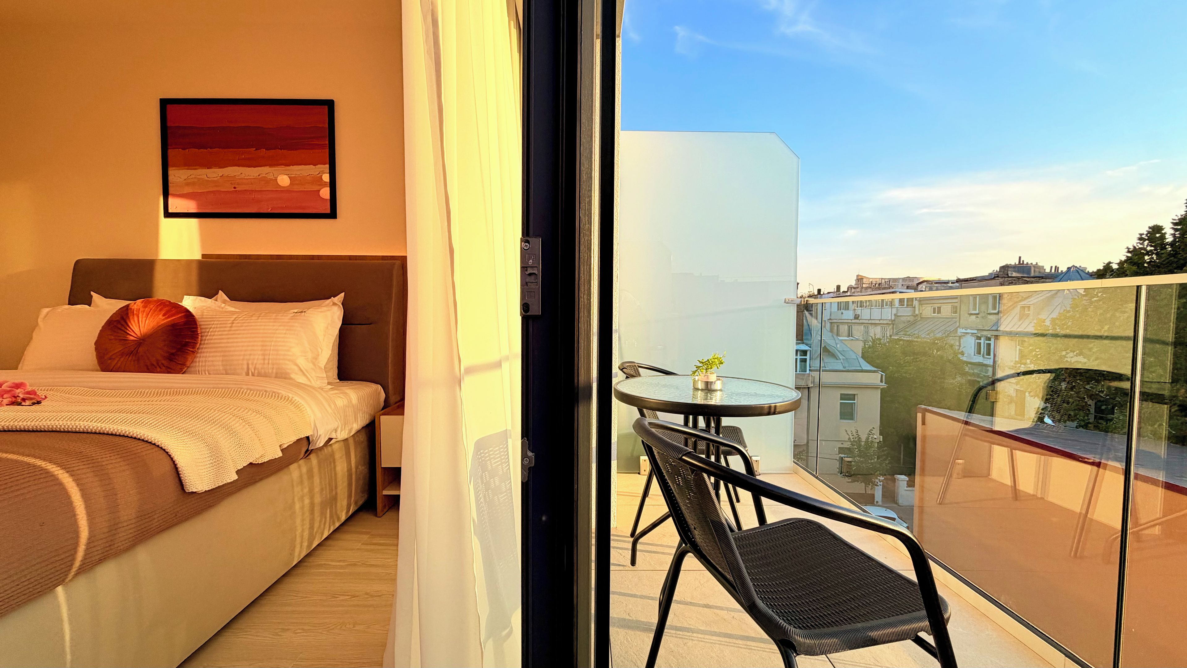 Habitación de hotel con cama, un pequeño balcón con mesa y silla, y vistas a la ciudad.