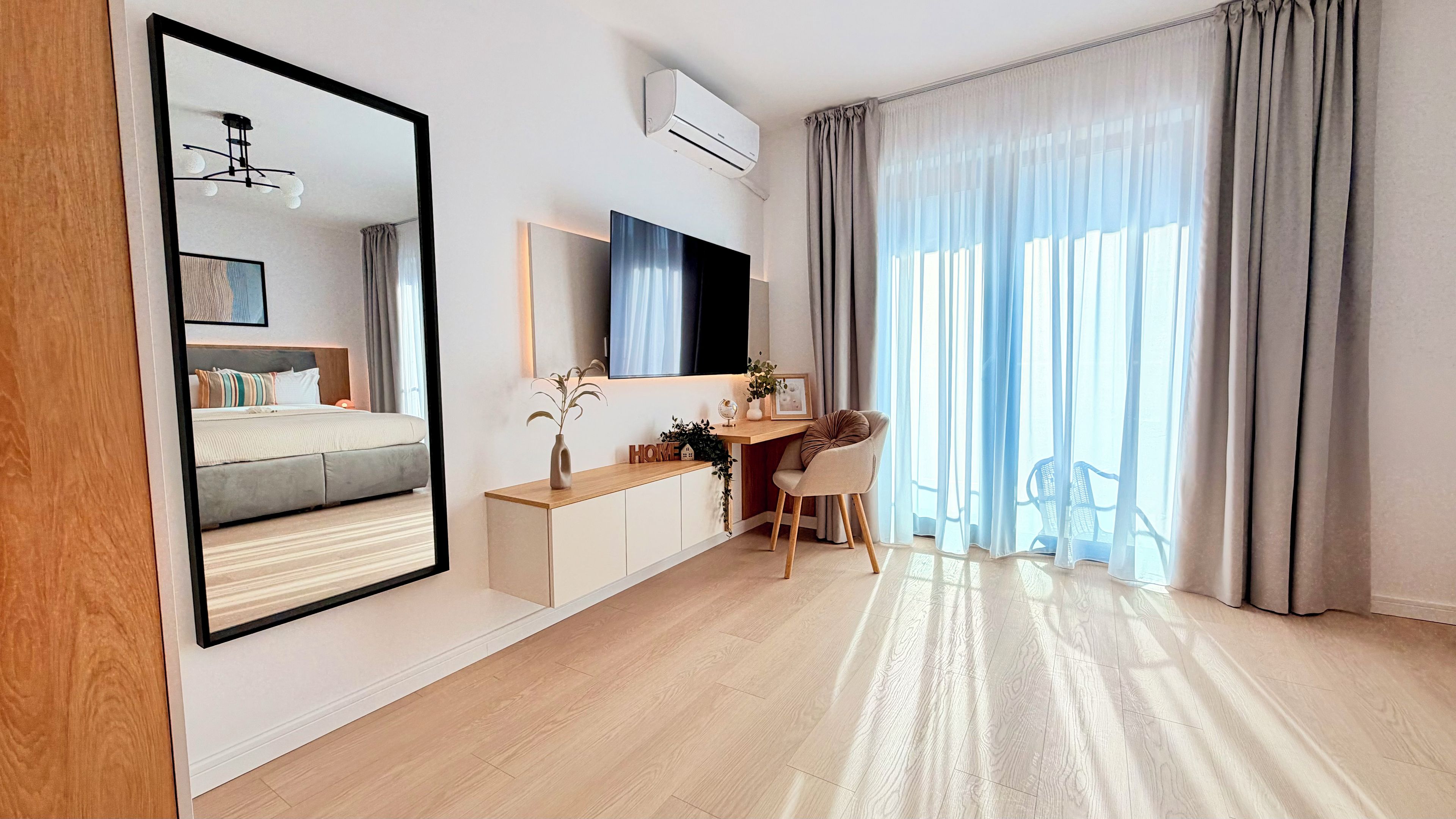 A modern bedroom with a large mirror, a flat-screen TV, a desk with a chair, and a large window with curtains.