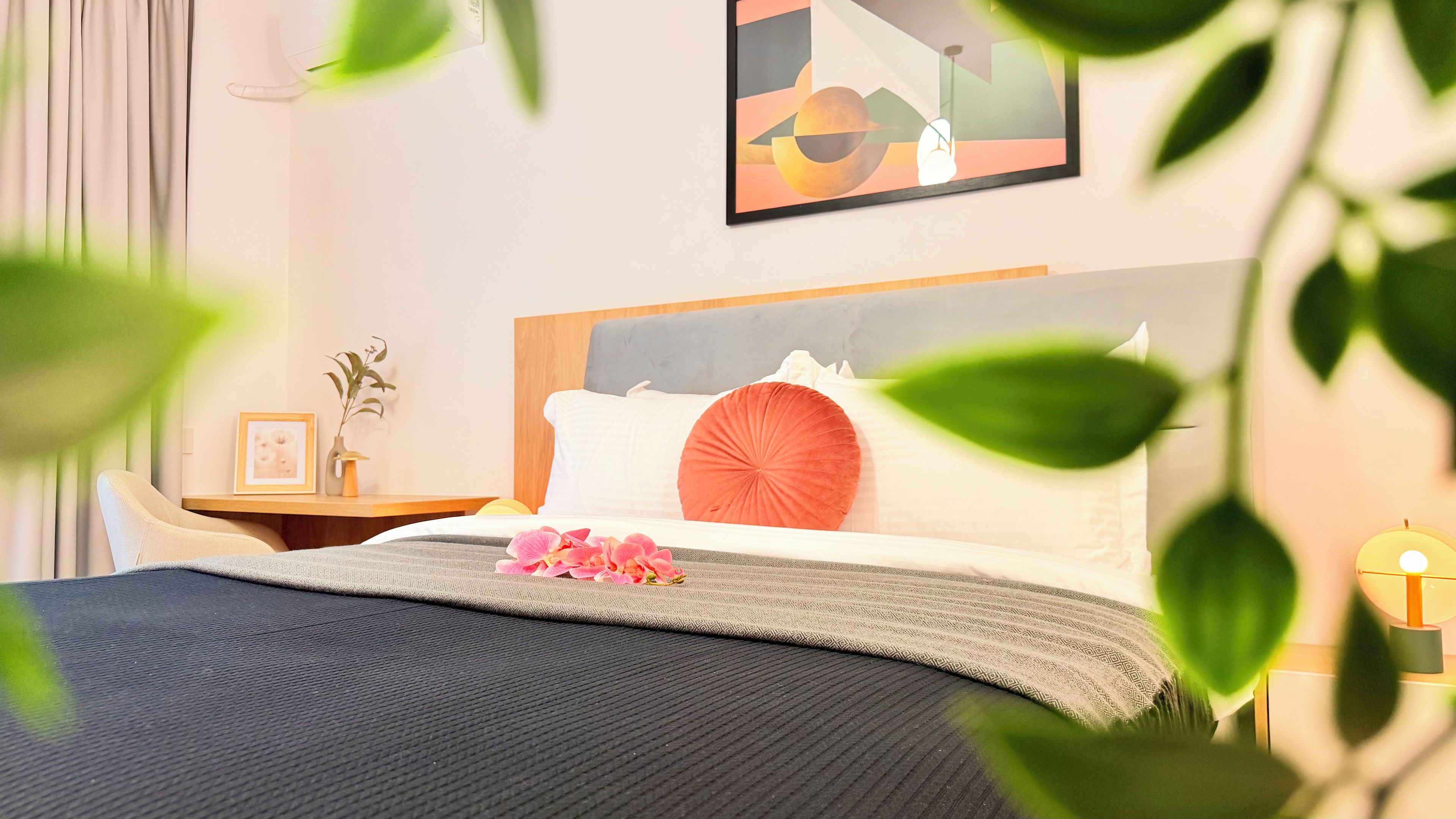 A neatly made bed with a pillow, a bedside table with a vase and picture frame, and a wall with a framed abstract painting.