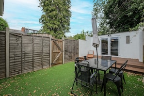 Un jardin avec une clôture en bois, une table en verre avec des chaises et un bâtiment blanc avec une porte.