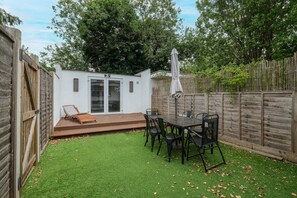 Un jardin avec une terrasse en bois, une table entourée de chaises et un bâtiment blanc doté de portes vitrées.
