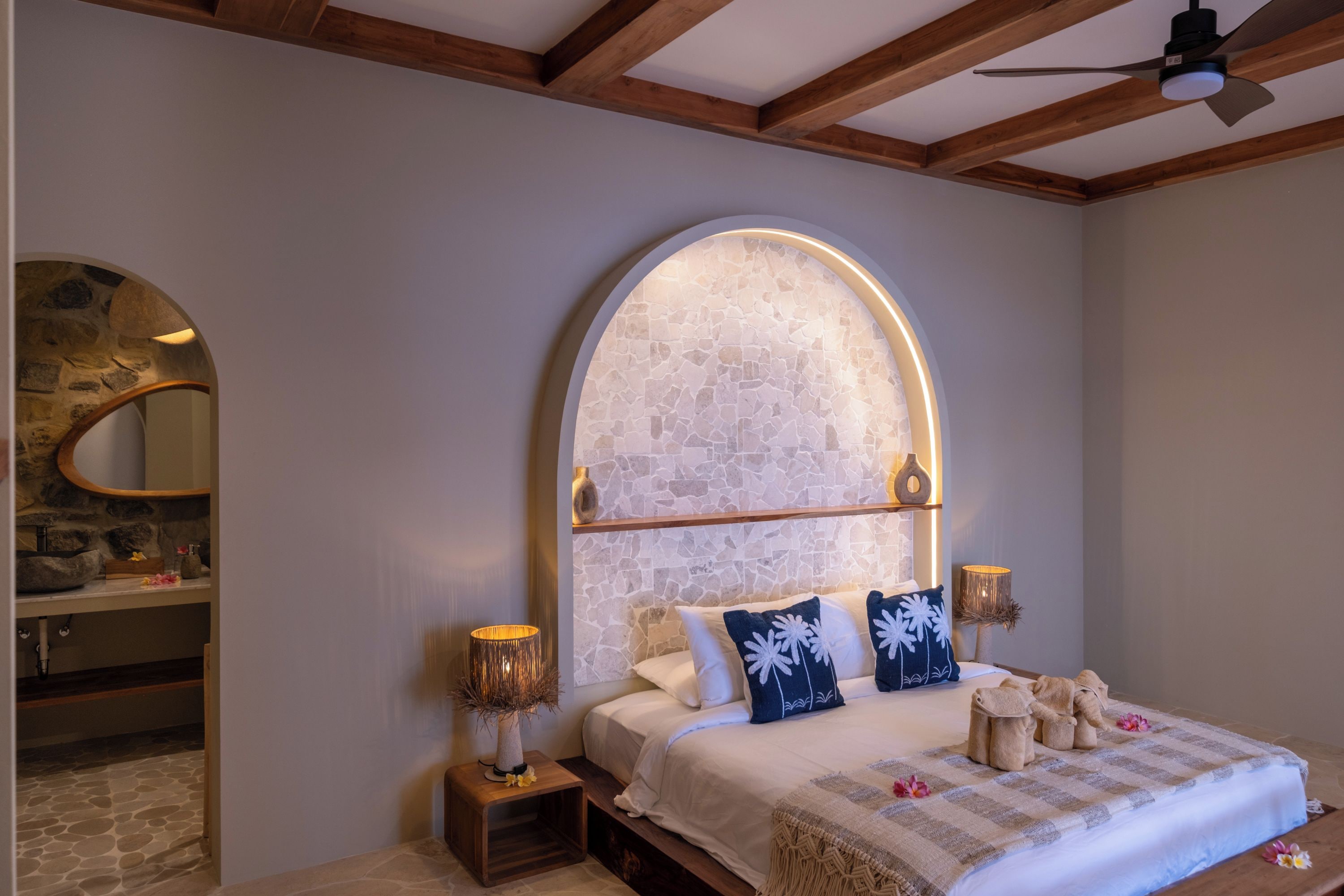 Un dormitorio con arco de pared de piedra, una cama con ropa blanca y una mesita de noche con lámpara.