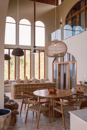 A spacious room with a high ceiling, large windows, a round wooden table, wicker furniture, and a hanging light fixture.