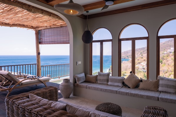A room with a view of the sea, a balcony with lounge chairs, and a seating area with cushions.