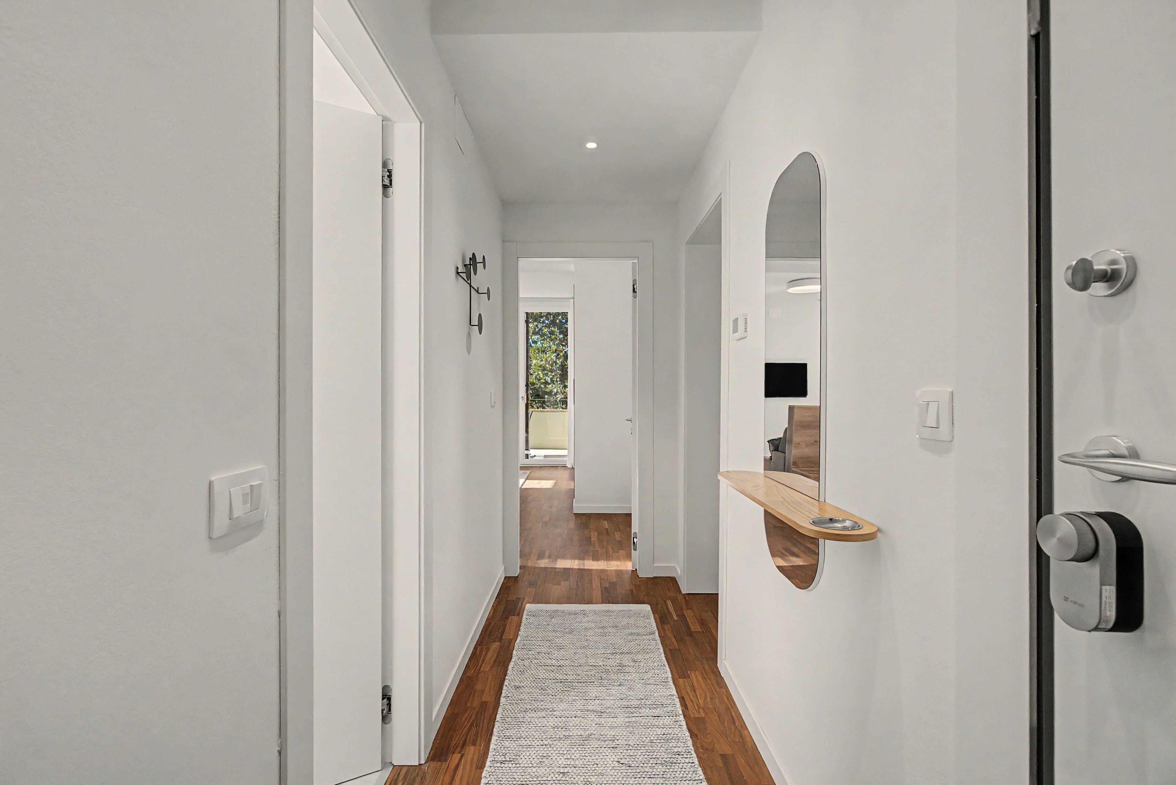 A hallway with white walls, wooden flooring, and a rug leading to a room with a mirror and a wooden shelf.