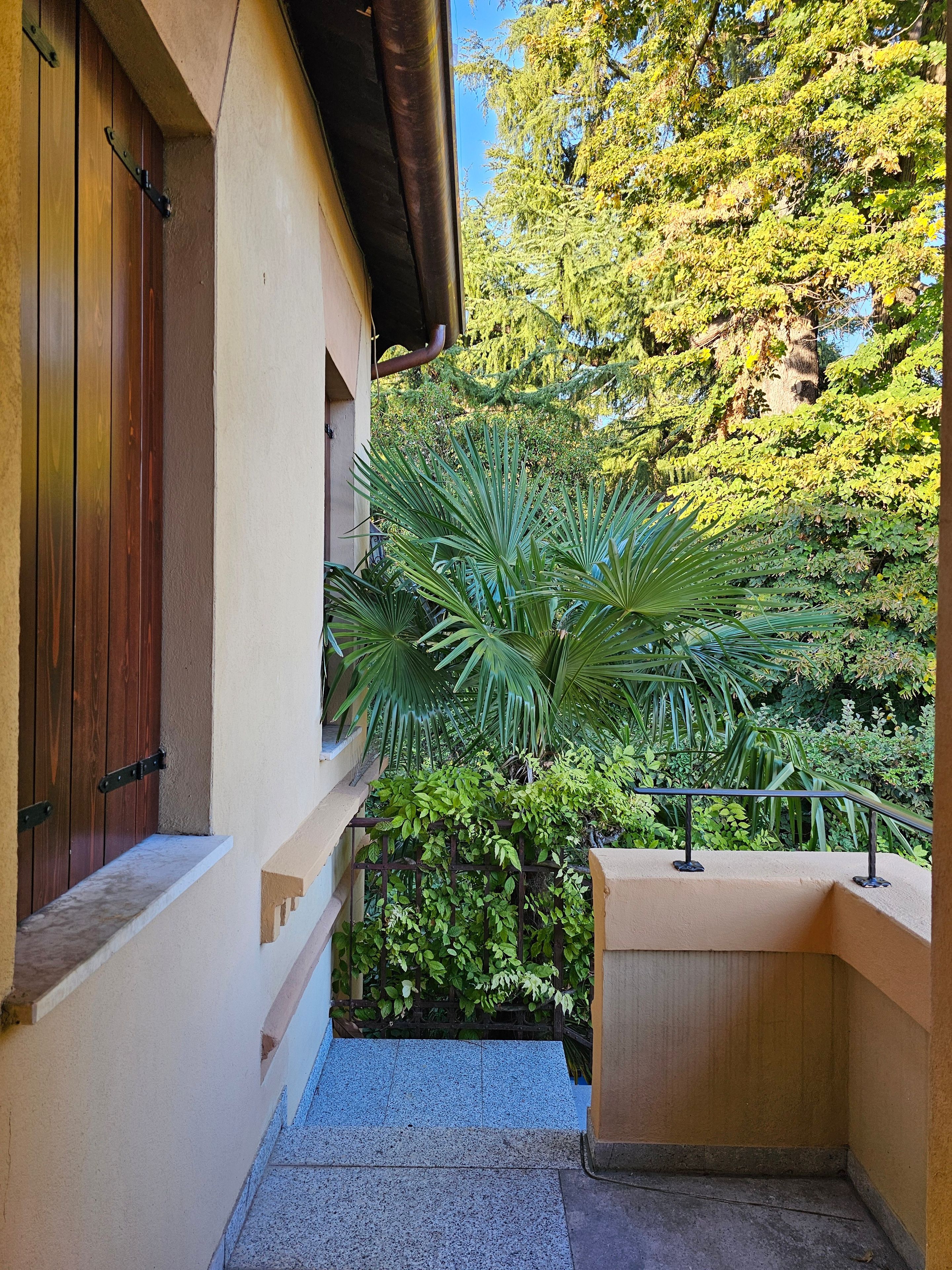 A narrow outdoor corridor with a wooden door and a metal railing overlooking a lush garden.