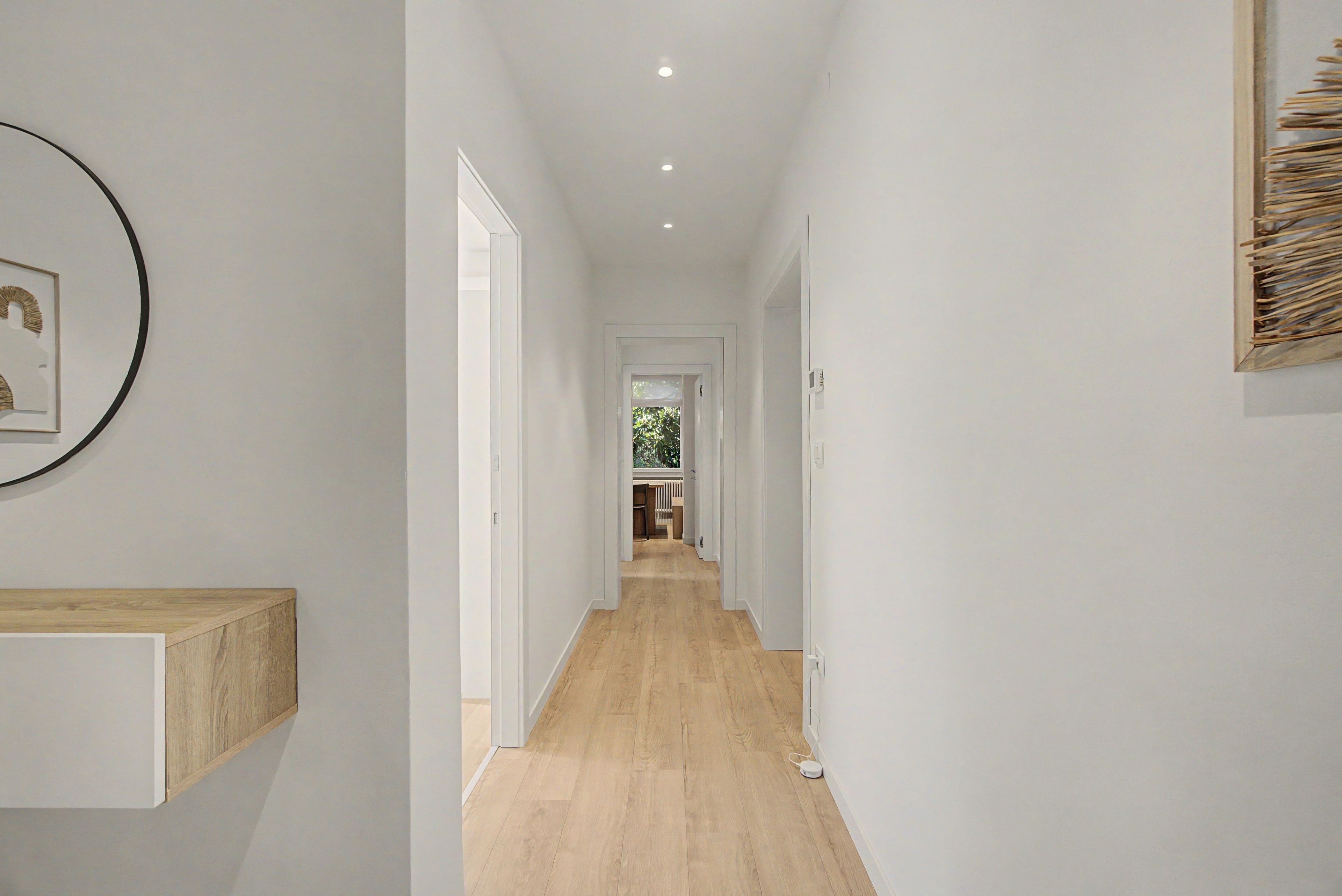 A hallway with a wooden floor, white walls, and a door leading to a room with a view of greenery.