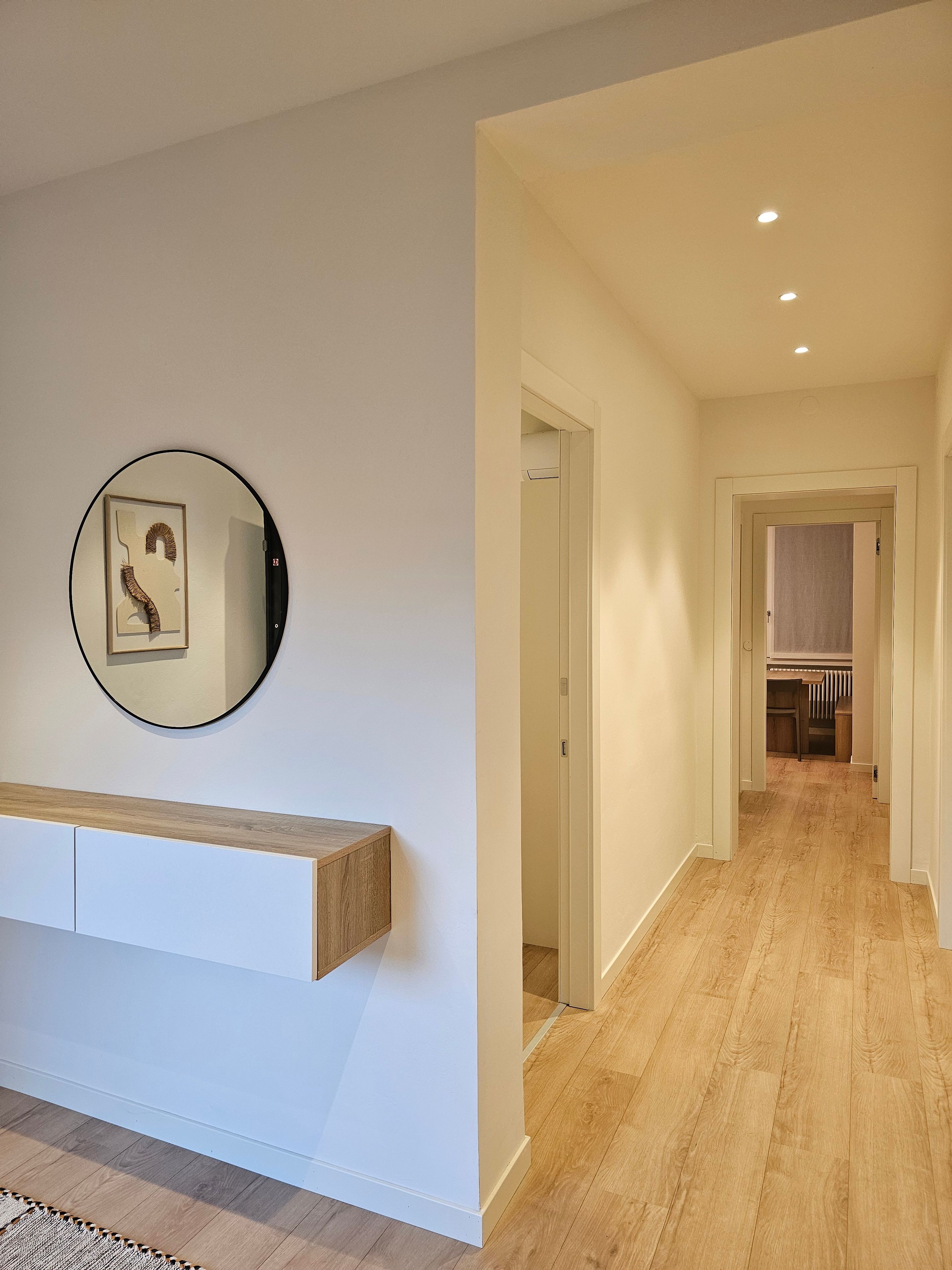 A hallway with a wooden floor, a white wall, and a framed artwork above a wooden bench.
