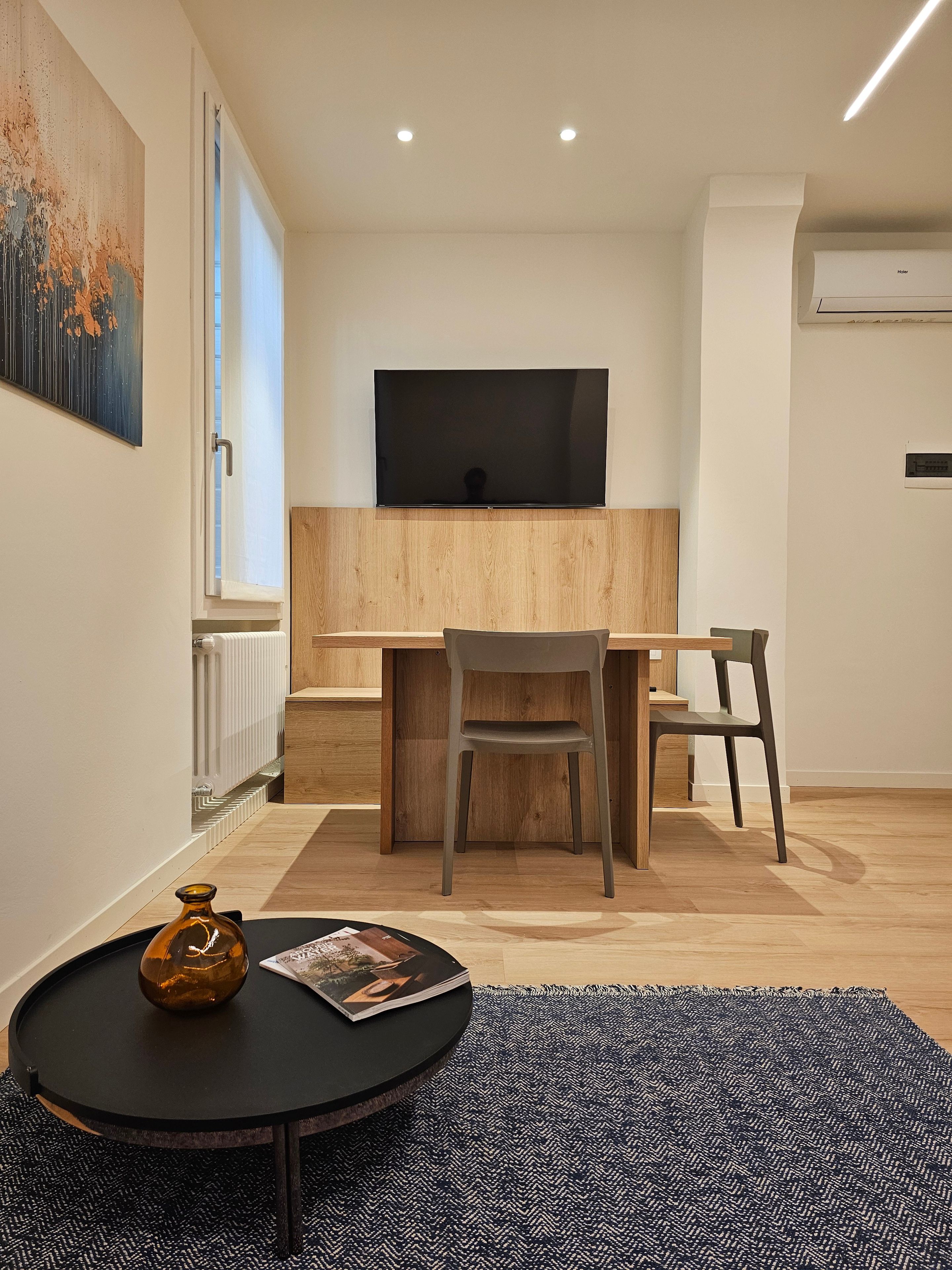 Ein modernes Zimmer mit einem Wand-Fernseher, einem Holztisch mit Stuhl, einem runden Tisch mit Flasche und Zeitschrift sowie einem gemusterten Teppich.