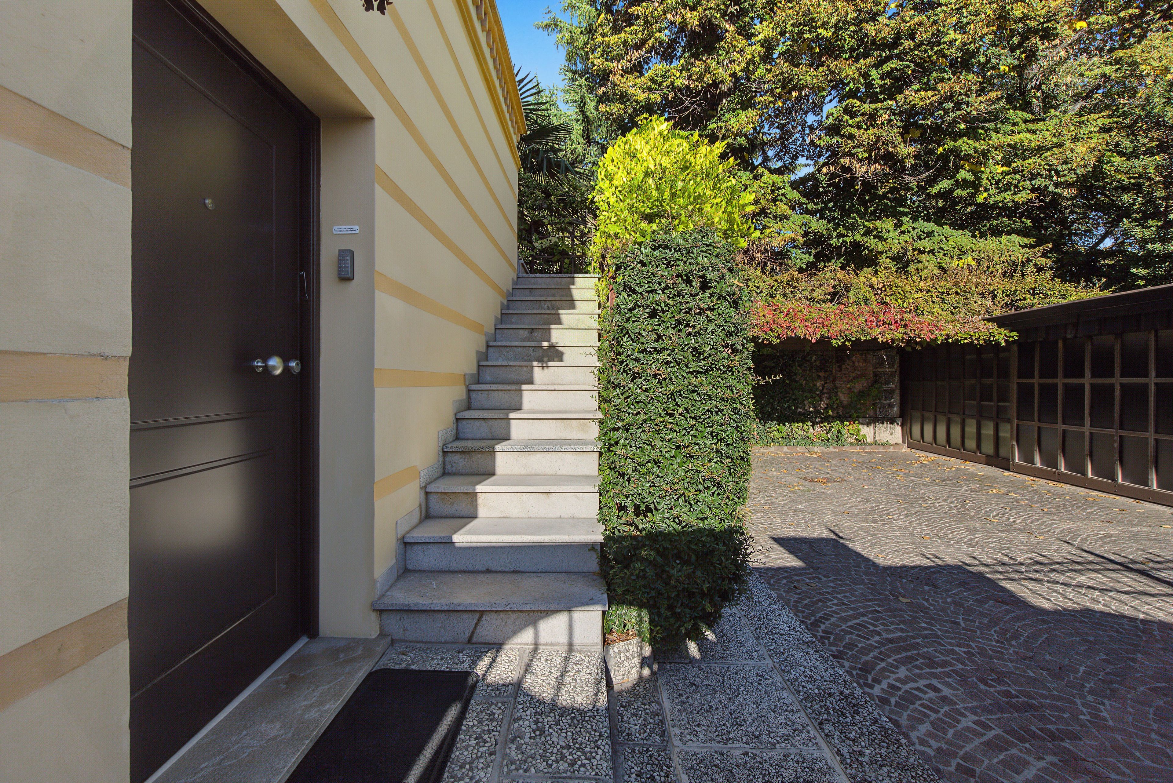 L’entrée d’un immeuble se caractérise par une porte sombre, un escalier menant à un palier, et un arbuste taillé avec soin.