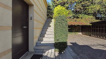 L’entrée d’un immeuble se caractérise par une porte sombre, un escalier menant à un palier, et un arbuste taillé avec soin.