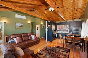 A spacious living room with a leather sofa, a dining area with wooden chairs, and a kitchen with a black refrigerator.
