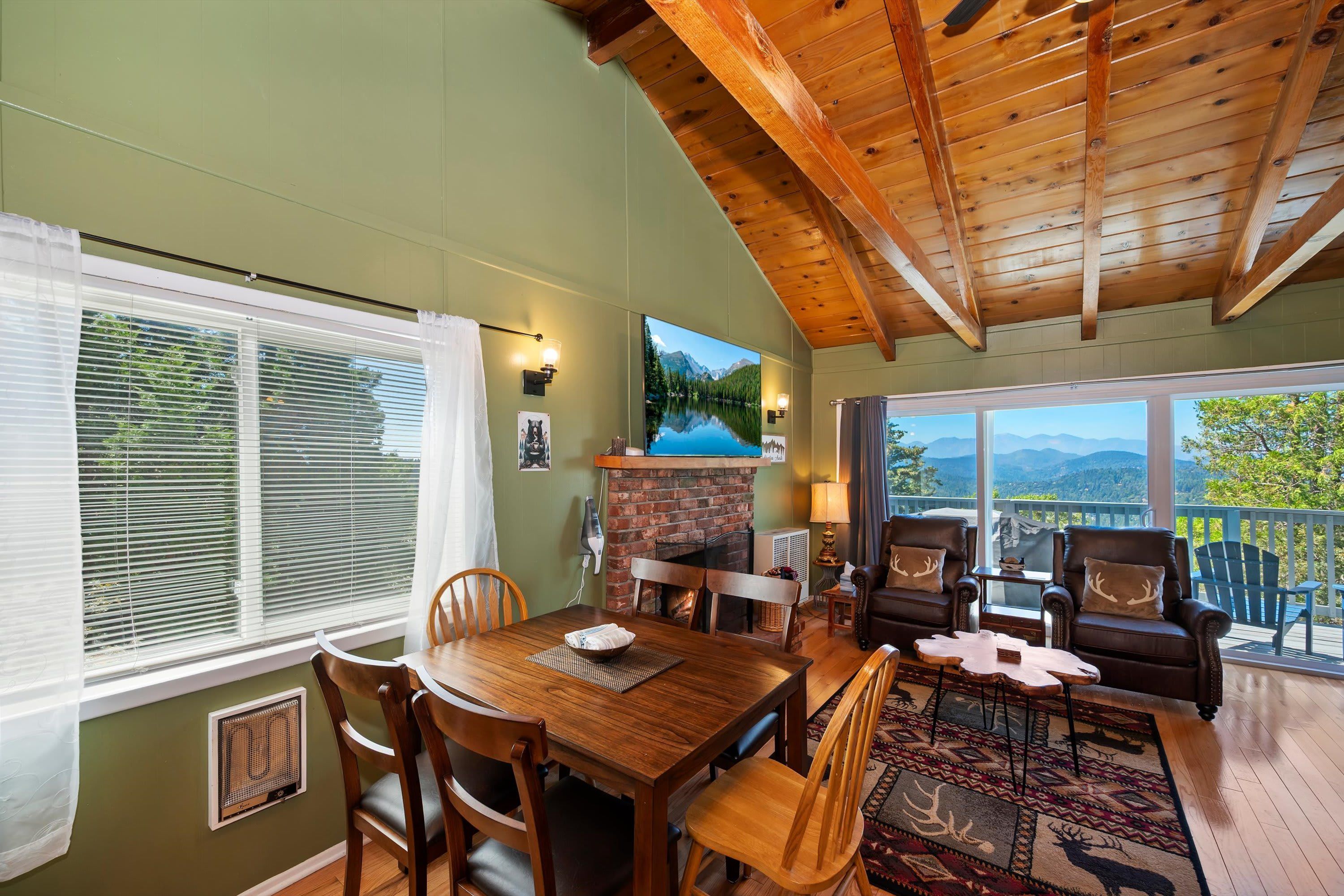 A cozy room with a wooden ceiling, a fireplace, a dining table with chairs, and a view of mountains through large windows.
