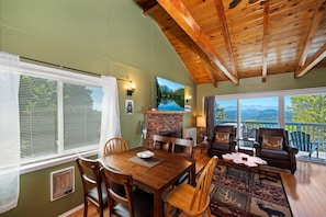 A cozy room with a wooden ceiling, a fireplace, a dining table with chairs, and a view of mountains through large windows.