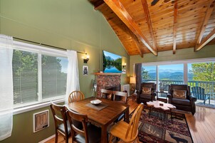 Une pièce chaleureuse avec un plafond en bois, une cheminée, une table à manger avec des chaises et une vue sur les montagnes par de grandes fenêtres.
