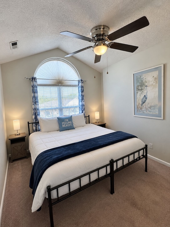 A bedroom with a bed, a ceiling fan, a window with curtains, a nightstand with a lamp, and a framed picture on the wall.