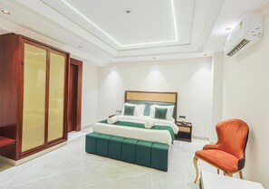 A modern bedroom with a large bed, a green upholstered bench, a wooden wardrobe, and an orange chair. - Clay Regency Hotel   (New Delhi)