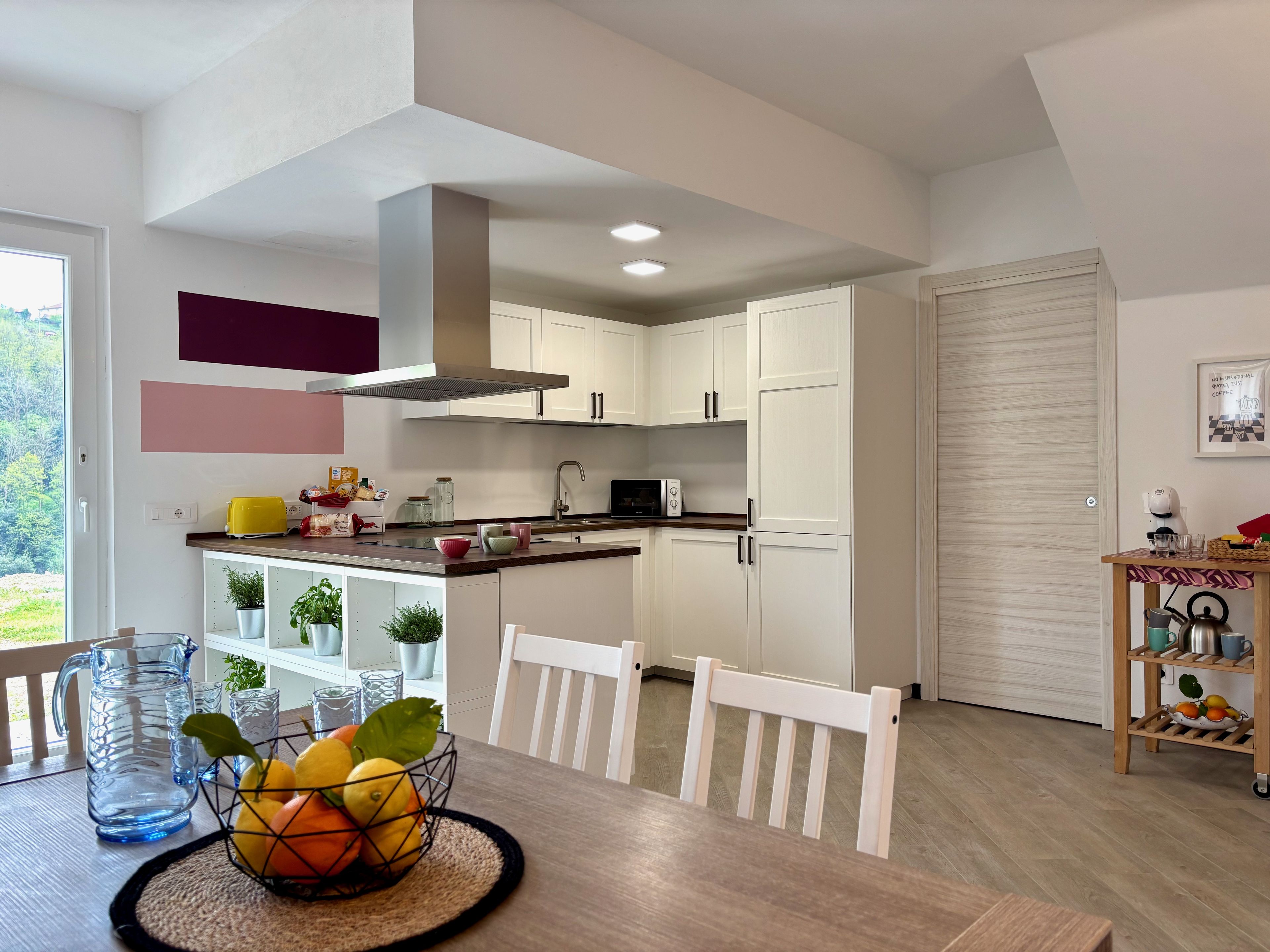A modern kitchen with white cabinets, a dining table with chairs, and a view of greenery outside.