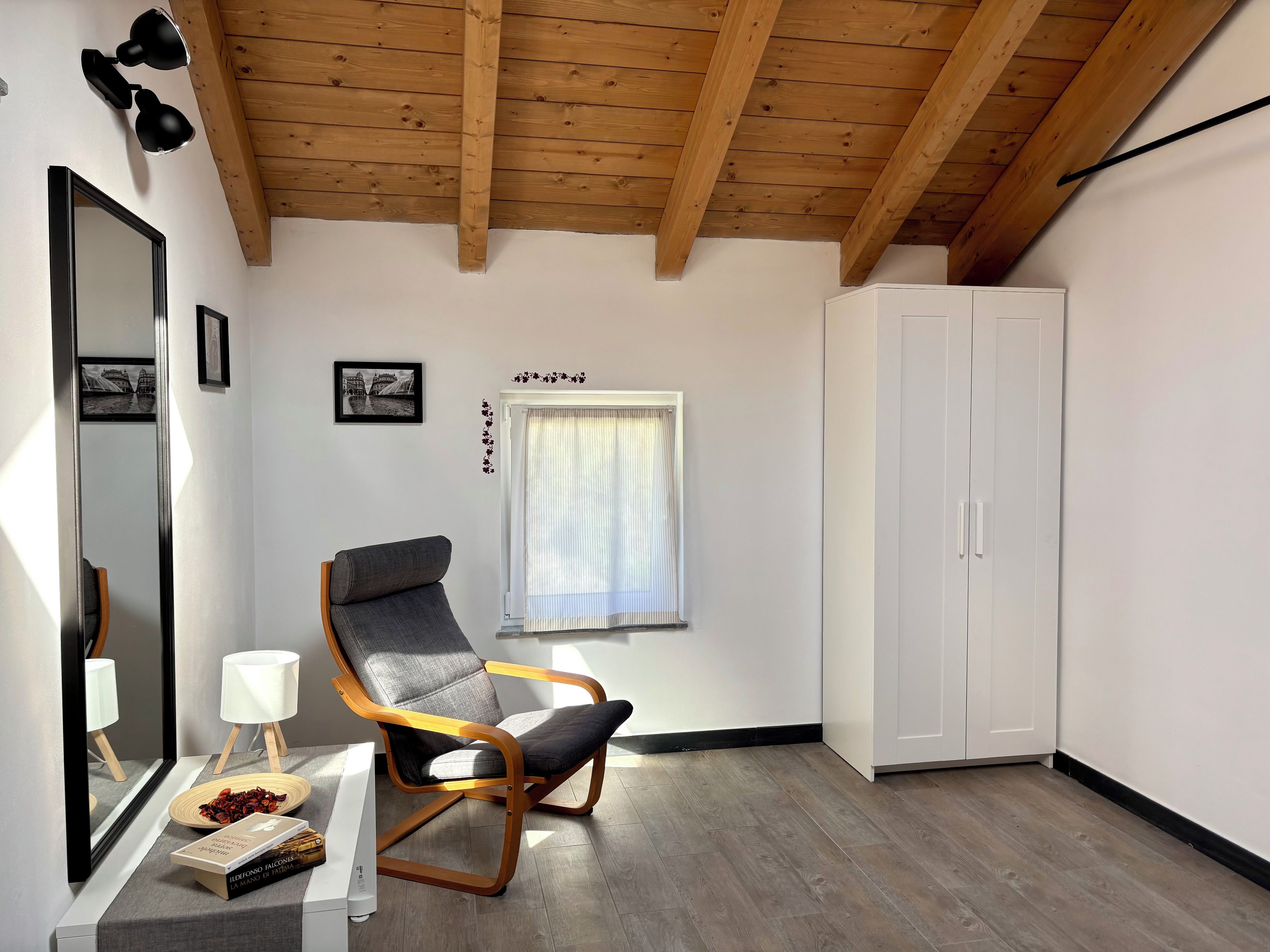 Une pièce avec un plafond en bois, une armoire blanche, une chaise grise, une petite table avec une lampe et une fenêtre avec des rideaux.