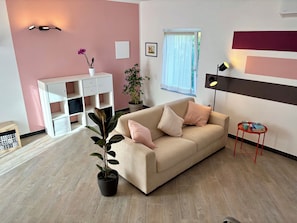 Una sala de estar con un sofá color beige, una planta en maceta, un armario blanco y una mesita auxiliar roja con libros.