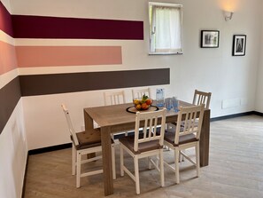 Comedor con mesa de madera, sillas blancas, frutero y ventana con persianas.