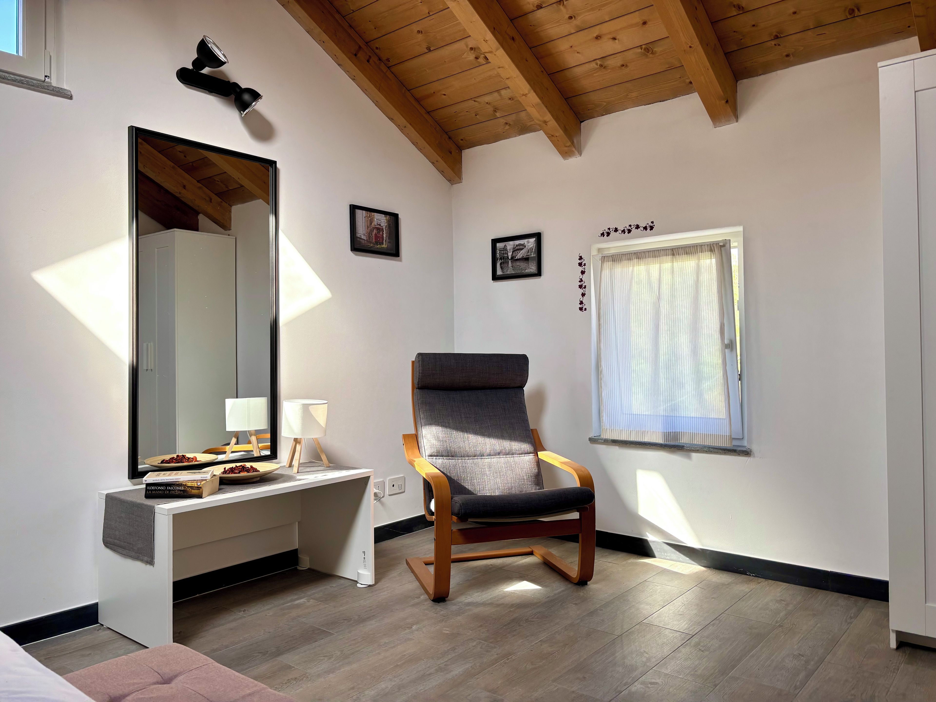 A room with a wooden ceiling, a desk with a mirror, a chair, and a window with a curtain.