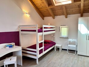 A bunk bed with a desk and chair, a small table, a cabinet, and a window with blinds.