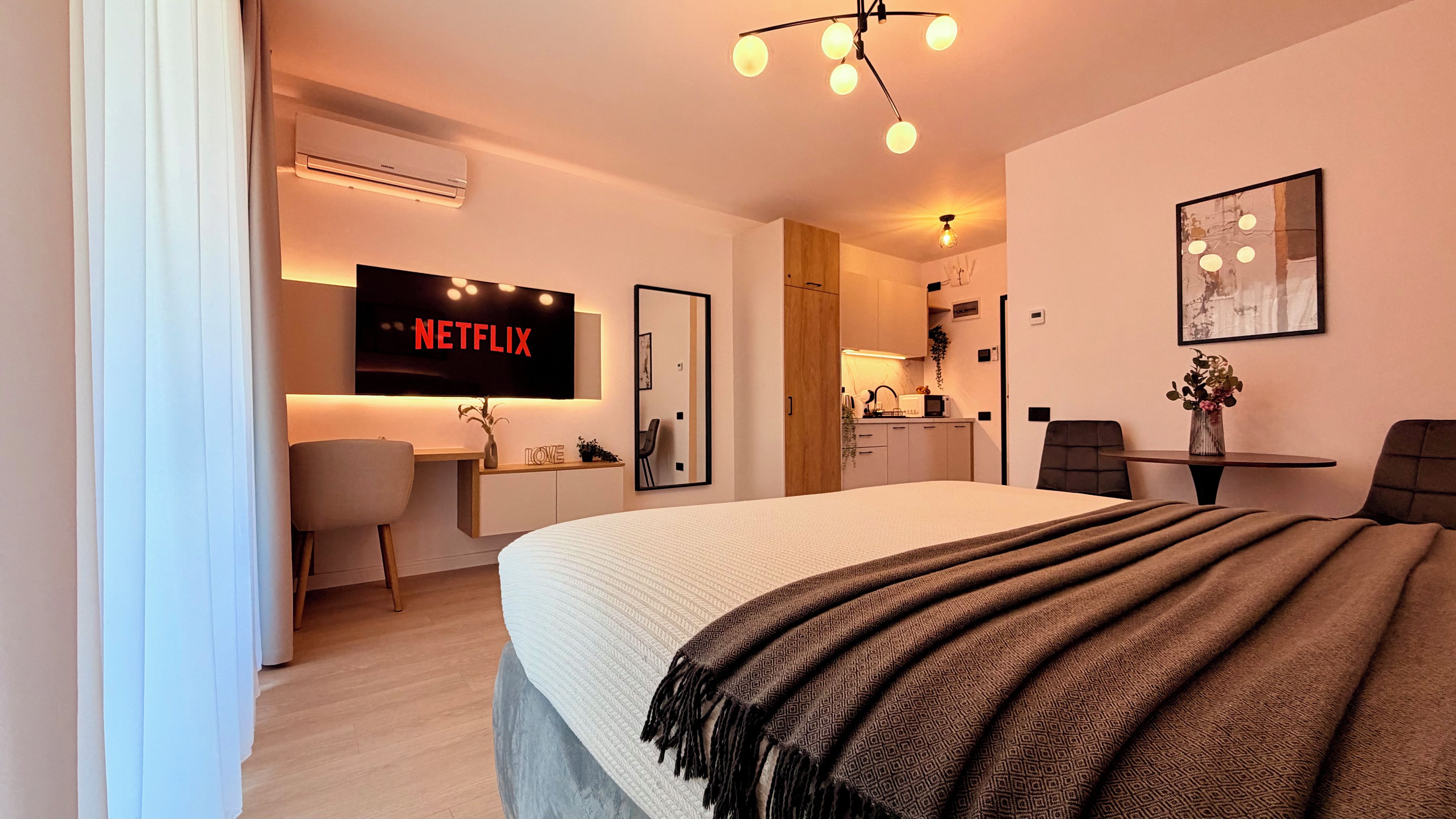 Una habitación de hotel moderna con una cama, un escritorio con una computadora, un televisor mostrando 'NETFLIX' y una kitchenette.