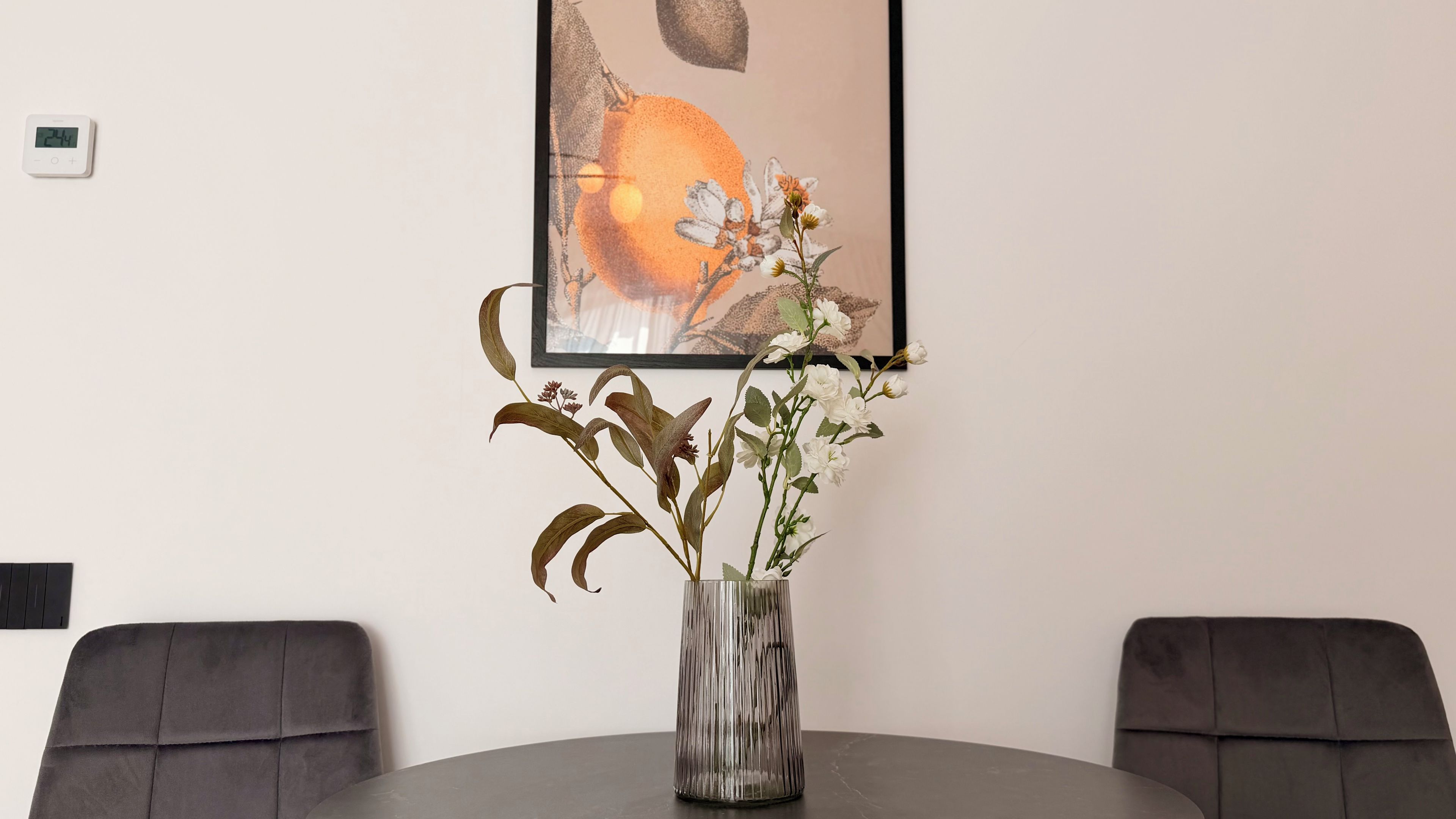 A dining table with a vase of flowers, two chairs, and a framed artwork on the wall.
