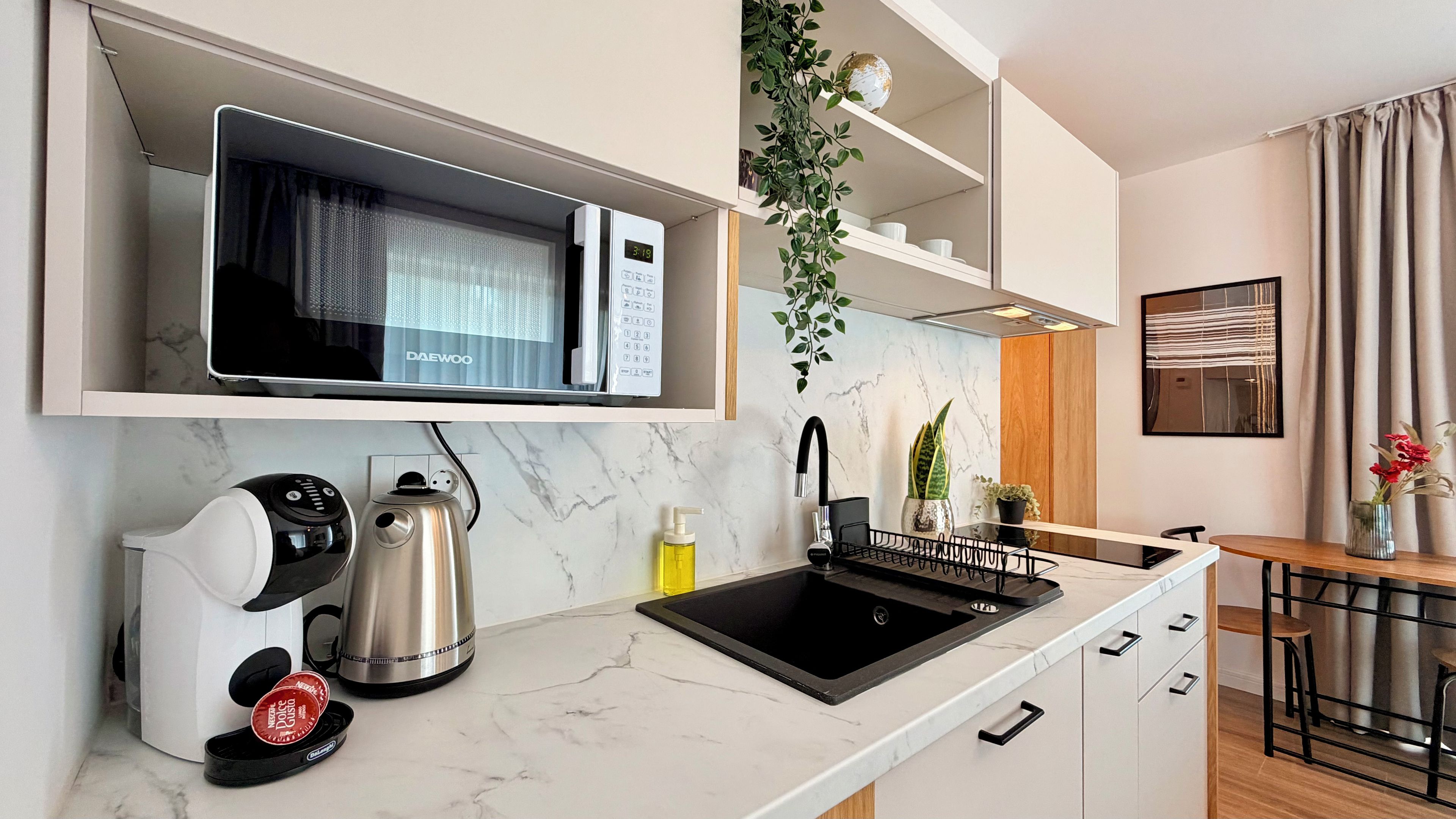 A modern kitchen with a built-in microwave, a kettle, a coffee machine, a sink, and a plant.